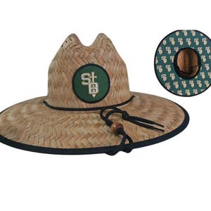 May include: A straw hat with a wide brim and a black trim. The hat features a green and white patch with the letters "SB". The hat has a black chin strap with a wooden bead. The underside of the brim has a green pattern.