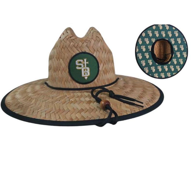 May include: A straw hat with a wide brim and a black trim. The hat features a green and white patch with the letters "SB". The hat has a black chin strap with a wooden bead. The underside of the brim has a green pattern.