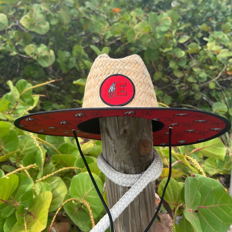 May include: A straw sun hat with a wide brim. The hat features a red band with a repeating pattern and a circular patch with a letter "E" and an image of a Native American headdress. Black chin strap.