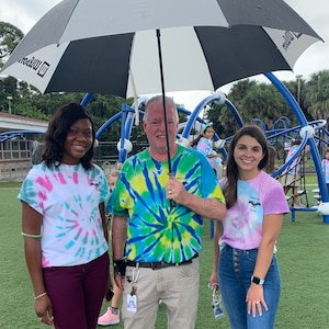 May include: Three people stand under a black and white umbrella on a playground. The person in the middle is wearing a green, blue, and yellow tie-dye shirt. The person on the left is wearing a pink and blue tie-dye shirt. The person on the right is wearing a pink and white tie-dye shirt.