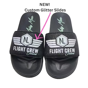 Può includere: Un paio di infradito nere glitterate con la scritta "FLIGHT CREW General" e un design ad ali bianche. Le infradito hanno la scritta "Molly Mae" sul plantare e la scritta "NEW! Custom Glitter Slides" in alto.