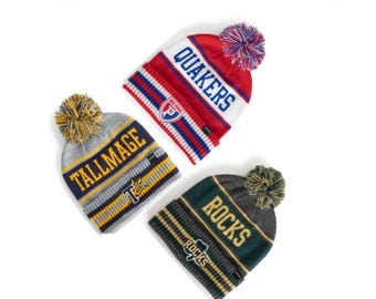 May include: Three knit beanies with pom-poms. One is red and white with "QUAKERS" in blue. Another is gray and yellow with "TALLMAGE" in navy. The third is green and gray with "ROCKS" in gold. All have striped bands.
