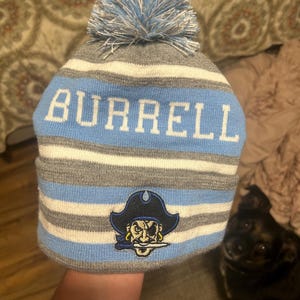 Pu&ograve; includere: Un berretto a maglia a righe grigie, bianche e azzurre, con la scritta "BURRELL" in lettere bianche. Il berretto presenta un pompon azzurro e una toppa con il logo di un pirata.