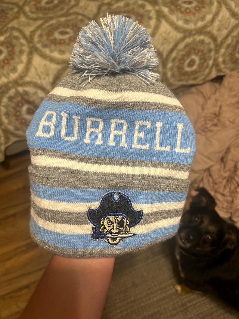 Pu&ograve; includere: Un berretto a maglia a righe grigie, bianche e azzurre, con la scritta "BURRELL" in lettere bianche. Il berretto presenta un pompon azzurro e una toppa con il logo di un pirata.