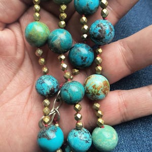 May include: A turquoise beaded necklace with gold-colored spacers. The beads are round and vary in shades of blue and green, with some showing brown marbling. The necklace is held in a hand.
