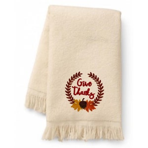 May include: A cream-colored kitchen towel with a fringed edge features an embroidered wreath with the words "Give Thanks" in red lettering. The wreath is decorated with fall-colored leaves and an acorn.