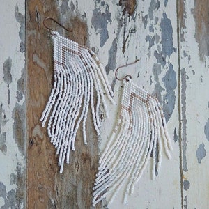 May include: White beaded earrings with a geometric pattern and fringe. The earrings are made with small white beads and have a brown wire hook.