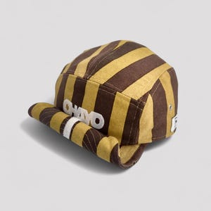 Puede incluir: Gorra a rayas marrones y amarillas con la palabra "CHAVO" bordada en blanco en la visera. La gorra tiene una visera plana y una corona redondeada. La gorra está hecha de una tela texturizada.