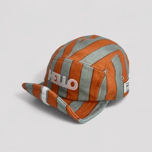 Puede incluir: Gorra de cinco paneles a rayas naranjas y grises con la palabra "HELLO" bordada en blanco. Tiene una visera corta y plana y una pequeña etiqueta en el lateral. Las rayas son verticales.