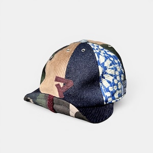 Puede incluir: Una gorra de béisbol de varios paneles con un diseño de patchwork que combina estampado de camuflaje, pana color canela, mezclilla azul oscuro y una tela acolchada con flores azules y blancas. Un parche de letra granate está cosido.