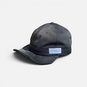 May include: A dark charcoal grey baseball cap with a short, curved brim and a structured crown. A light blue rectangular patch is stitched onto the front band, featuring the word Makshy written in a hand-drawn style with a black ink outline.