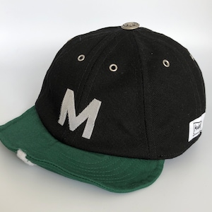 Black cotton cap with flat soft green cotton visor, metal eyelets on each panel, small velcro element on the middle of the visor, M letter velcro motif on front, Makshy label on side, and white studio background.