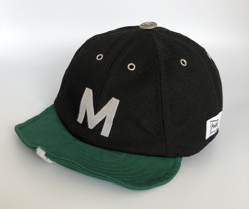 Black cotton cap with flat soft green cotton visor, metal eyelets on each panel, small velcro element on the middle of the visor, M letter velcro motif on front, Makshy label on side, and white studio background.