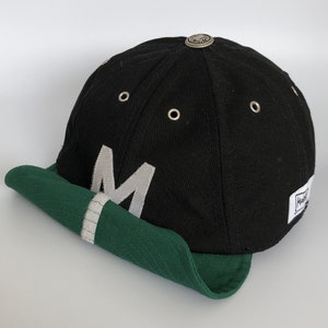 Black cotton cap with rolled up soft green cotton visor attached to the velcro motif. metal eyelets on each panel, small velcro element on the middle of the visor, M letter velcro motif on front, Makshy label on side, and white studio background.