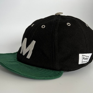 Black cotton cap with curved soft green cotton visor, metal eyelets on each panel, small velcro element on the middle of the visor, M letter velcro motif on front, Makshy label on side, and white studio background.