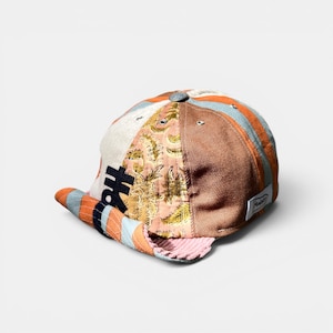 Puede incluir: Una gorra de varios paneles con una mezcla de telas que incluye sarga marrón, una sección a rayas azul claro y naranja, y un panel con estampado floral. La gorra tiene una visera corta y levantada con forro de pana rosa y una etiqueta blanca.