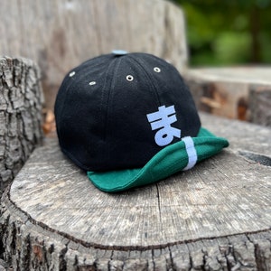 Black 8-panel baseball cap with a hiragana ま velcro letter emblem, green soft curved peak, and vintage-style metal squatchee. Cap is shown resting on a tree trunk in a park.