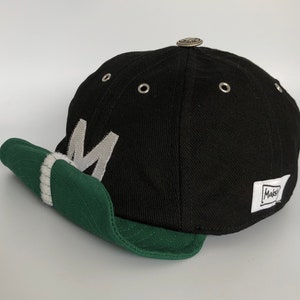 Black cotton cap with curved soft green cotton visor, metal eyelets on each panel, small velcro element on the middle of the visor, M letter velcro motif on front, Makshy label on side, and white studio background.