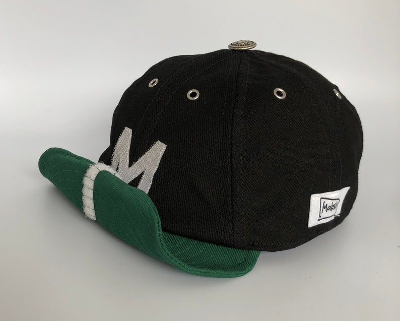 Black cotton cap with curved soft green cotton visor, metal eyelets on each panel, small velcro element on the middle of the visor, M letter velcro motif on front, Makshy label on side, and white studio background.