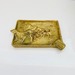 May include: A brass ashtray with a raised design of two dogs running. The ashtray is rectangular with rounded corners and a slightly curved edge.