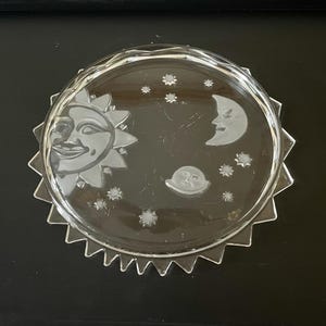 May include: A clear glass plate with a sun, moon, stars, and a jagged edge design. The sun has a smiling face. The moon and stars are etched into the glass. The plate is round and sits on a black surface.