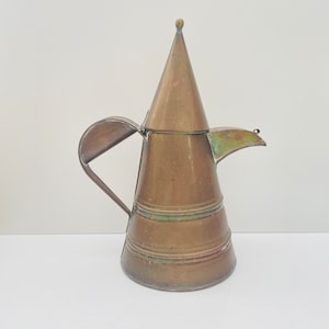 May include: A vintage copper pitcher with a conical lid and a curved handle. The pitcher has a spout and a series of horizontal bands around the body. The metal has a patina, showing signs of age and use.