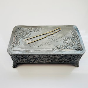 May include: A silver trinket box with an ornate design and the words "Woman's Friend" engraved on the top. A gold hair clip is resting on the box.