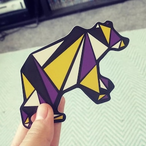 Puede incluir: Una pegatina de oso geométrico con un esquema de color morado, amarillo y negro. El oso está de pie con la cabeza girada hacia la derecha.