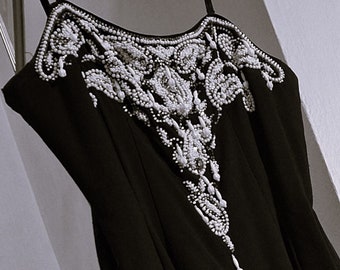 May include: A black sleeveless dress with a white beaded design on the neckline. The beads are arranged in a floral pattern.