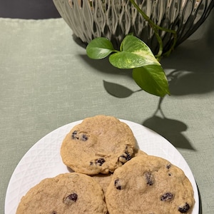 May include: A white plate holds three round, golden-brown cookies with dark raisins. The cookies are on a white plate with a textured pattern. A shiny, silver decorative vase and green plant are in the background.