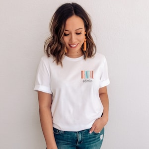 May include: A white t-shirt with the text "DENTAL admin" printed on the front in colorful letters. The shirt is being worn by a person with long brown hair and orange tassel earrings.
