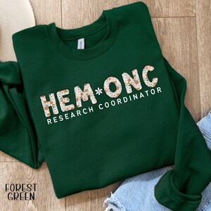 May include: A forest green sweatshirt with the text "HEM*ONC RESEARCH COORDINATOR" in white and floral print. The sweatshirt is folded on a wooden surface, with a tag that says "FOREST GREEN".