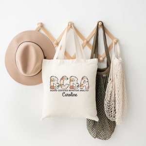 May include: A white canvas tote bag with the text "BCBA Board Certified Behavior Analyst Caroline" in black lettering. The letters are decorated with floral designs in shades of orange, yellow, and white.