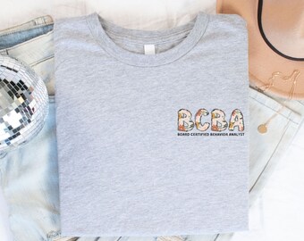 May include: A gray t-shirt with the text "BCBA Board Certified Behavior Analyst" in floral letters.
