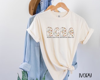 May include: Ivory t-shirt with a floral design that spells out "BCBA" and the text "Board Certified Behavior Analyst".