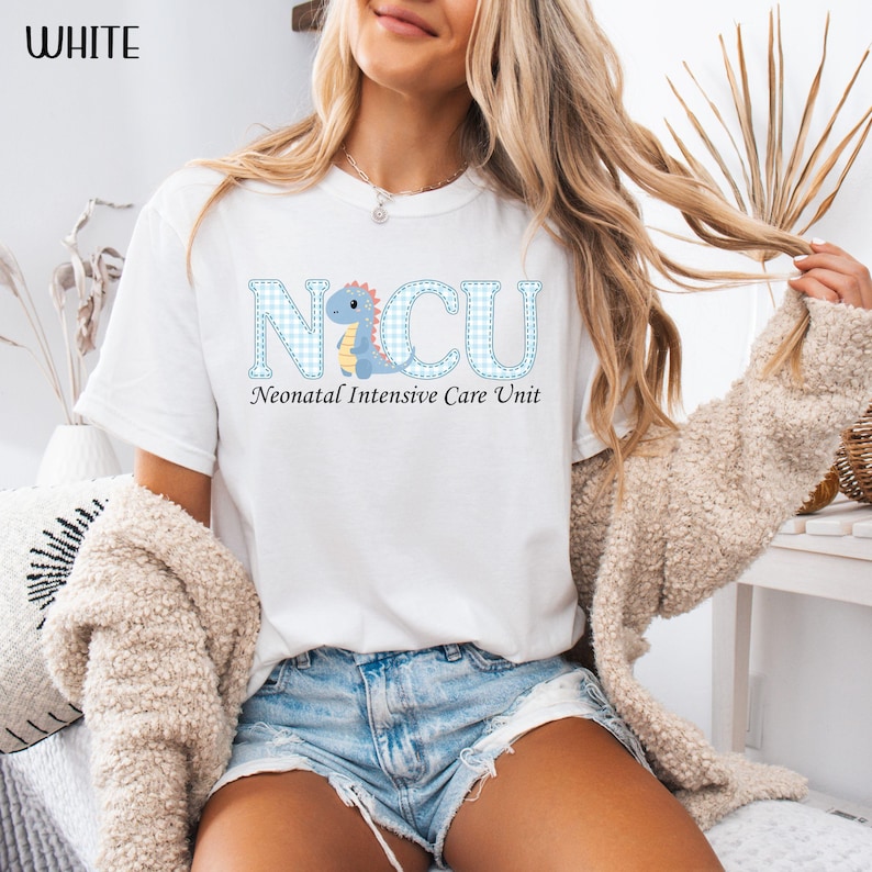 May include: White t-shirt with the text "NICU Neonatal Intensive Care Unit" in blue gingham letters and a cartoon dinosaur. The shirt is a crew neck style.