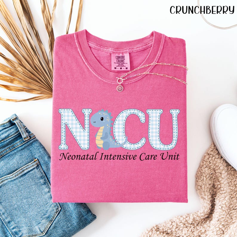 May include: A pink Comfort Colors t-shirt with the text "NICU Neonatal Intensive Care Unit". The letters are light blue with a gingham pattern and a cartoon dinosaur. A gold necklace is draped across the shirt.
