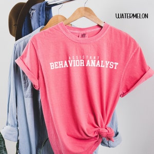 May include: A pink t-shirt with the text "Assistant Behavior Analyst" in white lettering.