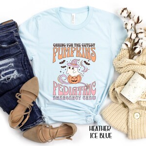 May include: A light blue t-shirt with a graphic of a ghost wearing a witch hat and sitting on a pumpkin. The text "Caring for the cutest pumpkins Pediatric Emergency Crew" is printed on the shirt.