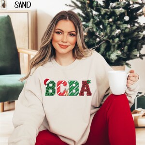 May include: A cream-colored sweatshirt with the text "BCBA" in festive Christmas-themed letters. The letters are decorated with green and red patterns, including a reindeer, holly, and a Santa hat.