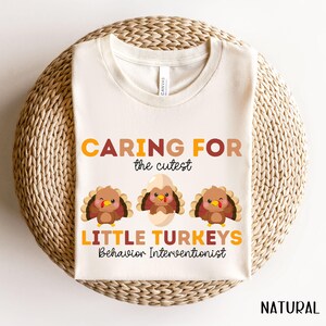 May include: A cream-colored t-shirt with the words "CARING FOR the cutest LITTLE TURKEYS Behavior Interventionist" in orange and brown. Three cartoon turkeys are below the text. The word "NATURAL" is at the bottom right.