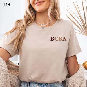 May include: Tan colored t-shirt with the text "BCBA Emily" in brown, red, and beige letters.