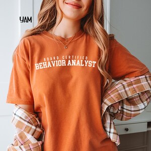 May include: An orange t-shirt with the text "BOARD CERTIFIED BEHAVIOR ANALYST" in white block letters. The shirt has short sleeves and a crew neck. A plaid shirt is draped over the shoulders.