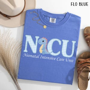 May include: A blue t-shirt with the text "NICU" in a white and blue gingham pattern, with a cartoon dinosaur illustration. The text "Neonatal Intensive Care Unit" is below the letters. The shirt is next to a hat, jewelry, and sandals.