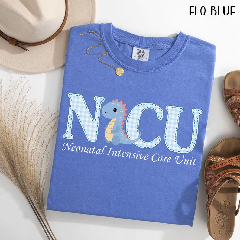May include: A blue t-shirt with the text "NICU" in a white and blue gingham pattern, with a cartoon dinosaur illustration. The text "Neonatal Intensive Care Unit" is below the letters. The shirt is next to a hat, jewelry, and sandals.