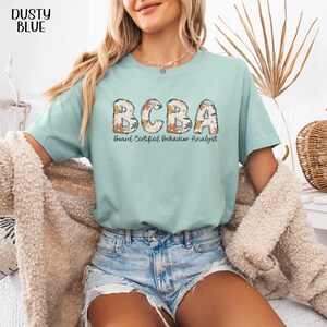 May include: A light blue t-shirt with the text "BCBA Board Certified Behavior Analyst" in floral letters.
