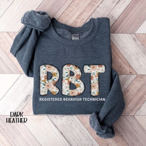 May include: Dark heather grey sweatshirt with the text "RBT" in large, white letters outlined in black. The letters are filled with a floral pattern. Below the letters is the text "Registered Behavior Technician".