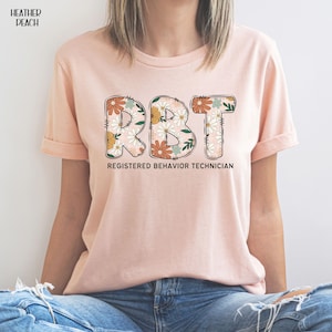 May include: A heather peach t-shirt with a floral design spelling out "RBT" and the text "Registered Behavior Technician".
