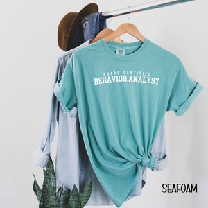 May include: A teal blue t-shirt with the text "Board Certified Behavior Analyst" in white lettering. The shirt is hanging on a hanger with a denim shirt and a brown hat in the background.