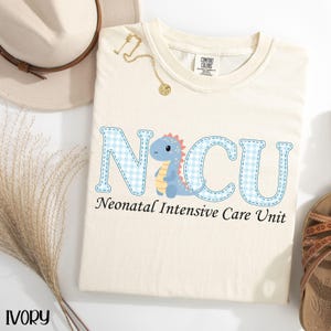 May include: Ivory-colored t-shirt with the letters "NICU" in light blue gingham print, featuring a cartoon dinosaur. The text "Neonatal Intensive Care Unit" is below the letters. A gold necklace and a hat are visible.